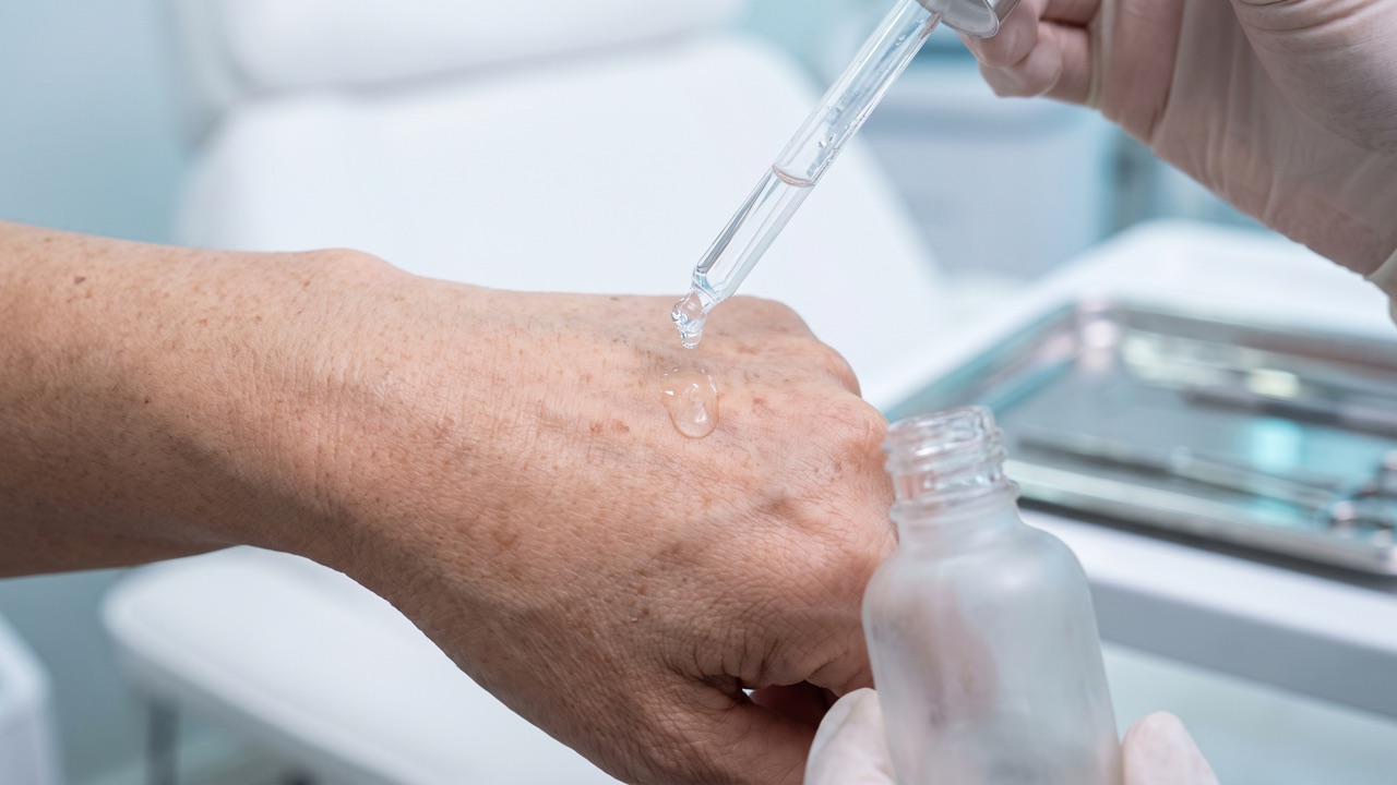 A glass dropper releasing a drop of clear serum onto the back of a mature hand in a clinic room