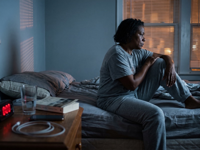 A woman sits on the edge of her bed in a dark bedroom at night, an alarm clock glowing on the nightstand